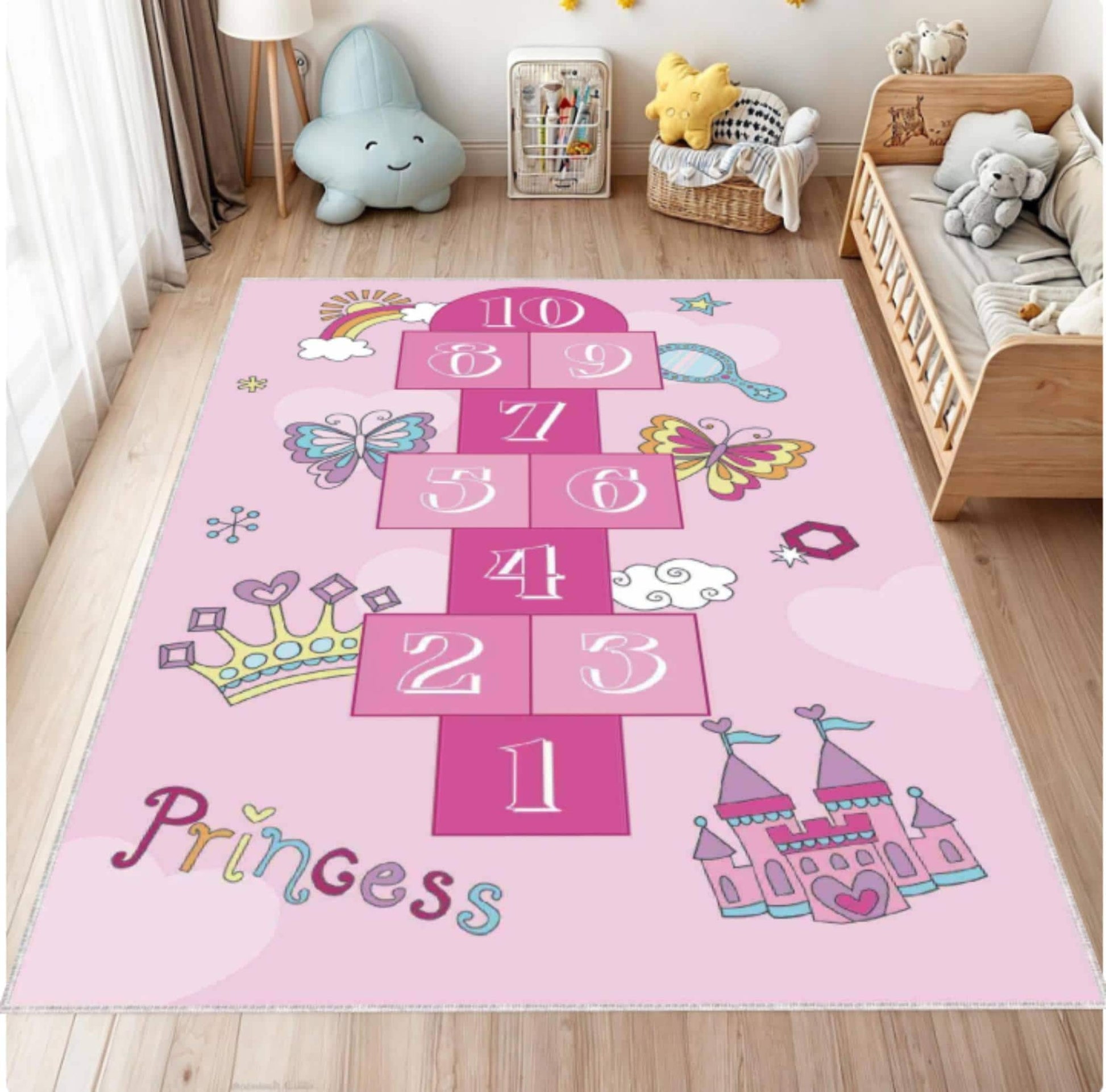 A pink children's area rug featuring a hopscotch grid, butterflies, crowns, a castle, the word 'Princess', and small decorative motifs, shown in a styled nursery with a wooden floor, crib, toys, and curtains.