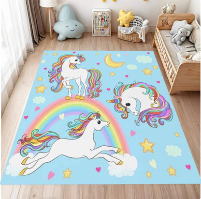 A rectangular children's room rug with a light blue background featuring two white unicorns, a rainbow, stars, hearts, and clouds. The rug is displayed on a light wood floor in a styled nursery setting with toys, a wooden crib, and a cloud-shaped lamp.