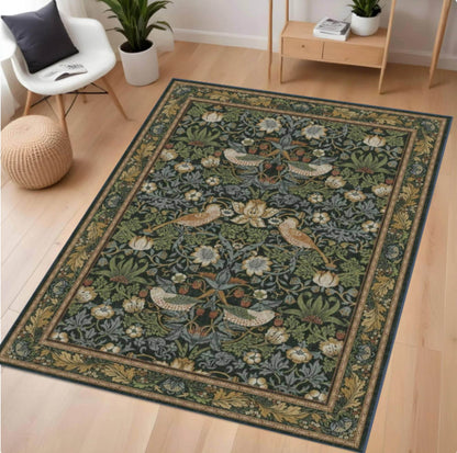 A rectangular William Morris area rug with a detailed botanical and bird pattern in muted green, beige, and blue-gray tones, shown on a light wood floor in a styled room with a chair, plant, and side table.