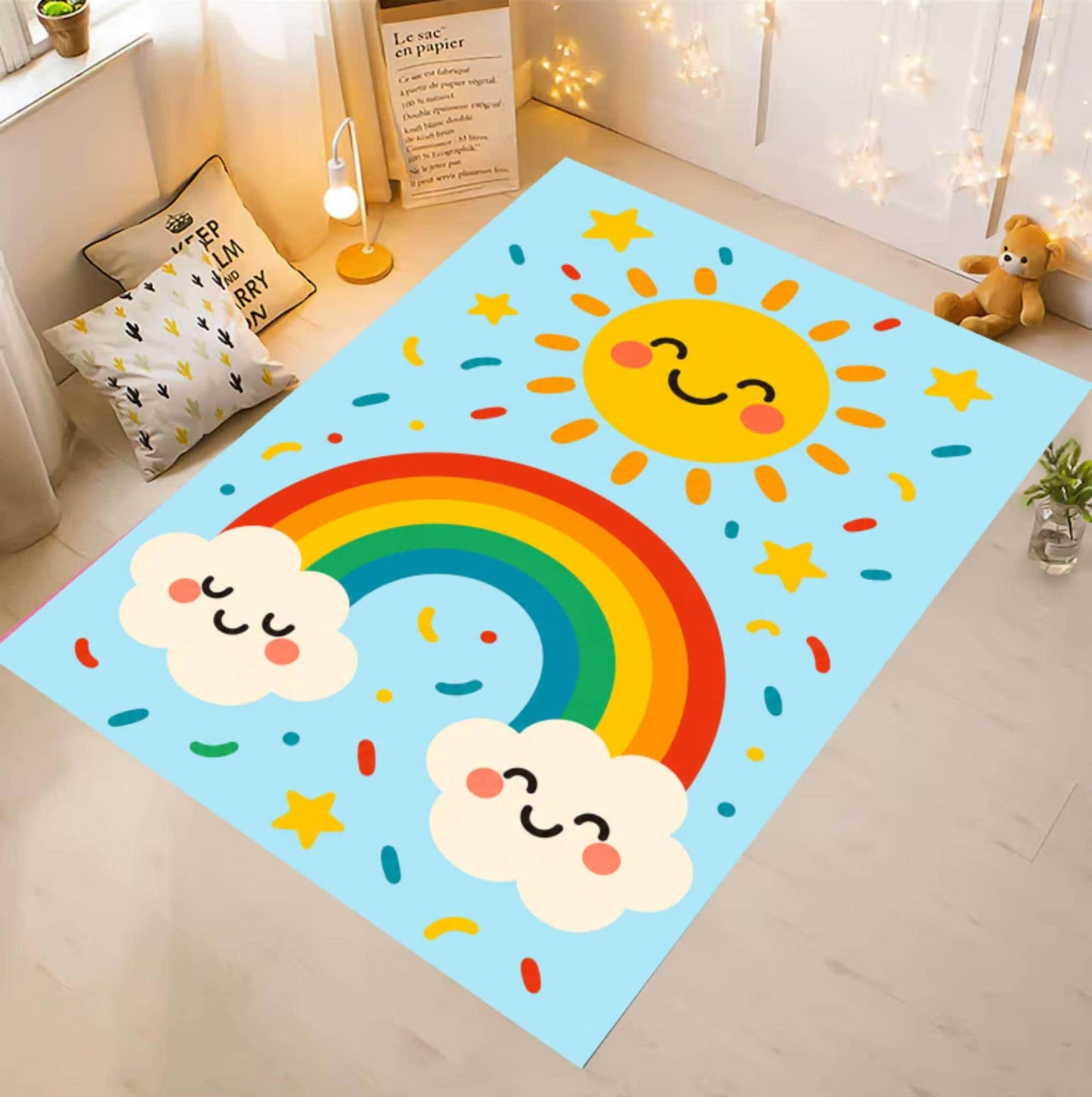 A rectangular baby play mat with a colorful cartoon design, shown in a styled nursery room setting. The mat features a light blue background with a smiling yellow sun, a rainbow, two white clouds with faces, and small stars and confetti.