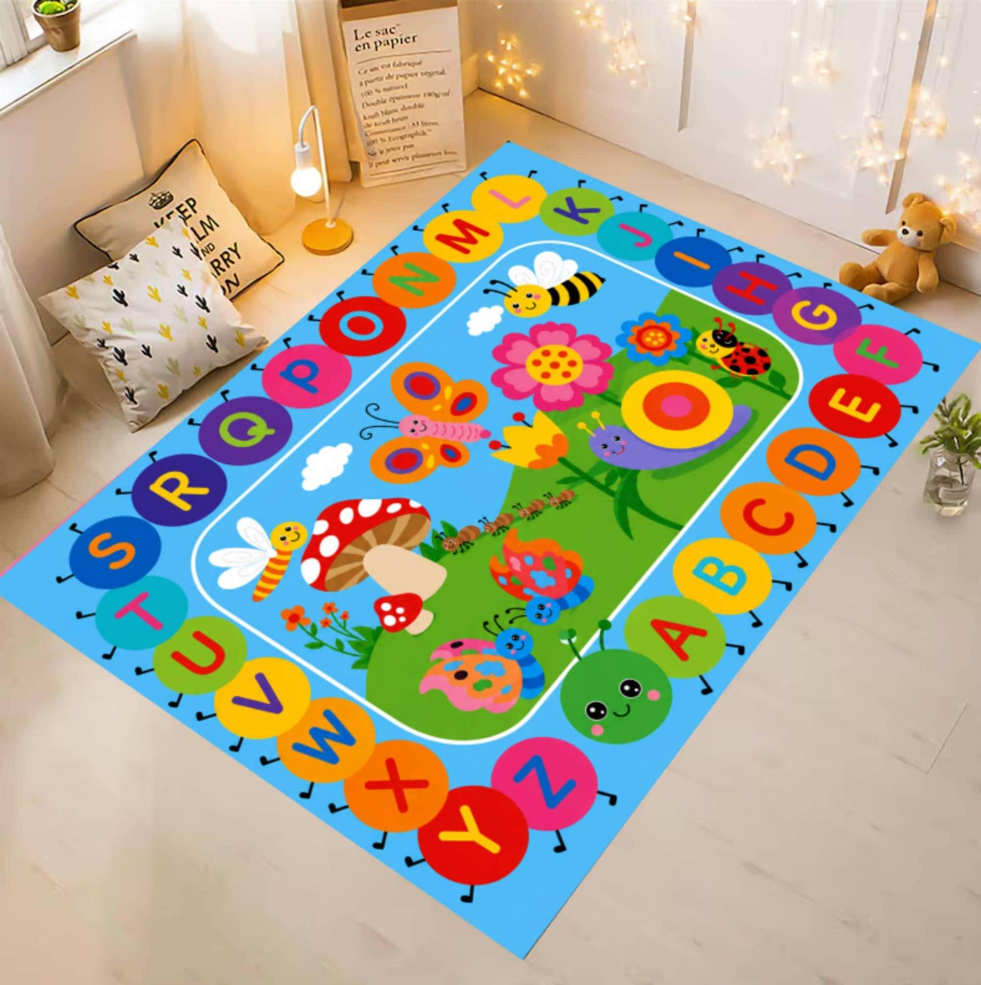A colorful children's alphabet play mat rug displayed on a light wood floor in a styled room. The rug features a bright blue border with alphabet letters in circular shapes and a green center depicting a whimsical garden with flowers, butterflies, and animals.