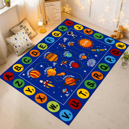 A colorful children's alphabet and space-themed area rug laid out on a light wood floor in a bright room. The rug has a blue background with a border of alphabet letters in colored circles and a central solar-system illustration including planets, stars, and a rocket.
