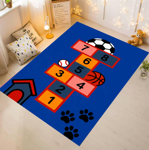 A rectangular blue pet-themed rug laid on a light wood floor in a cozy room. The rug features a colorful hopscotch-style number grid with soccer ball, baseball, basketball, and paw print graphics, plus a small dog house illustration.