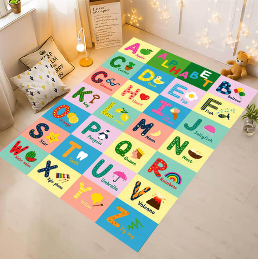 A colorful alphabet playmat laid out on a light wood floor in a child's room. The playmat features a grid of pastel-colored squares, each with a different uppercase alphabet letter, a cartoon illustration, and the letter's name in English and French.