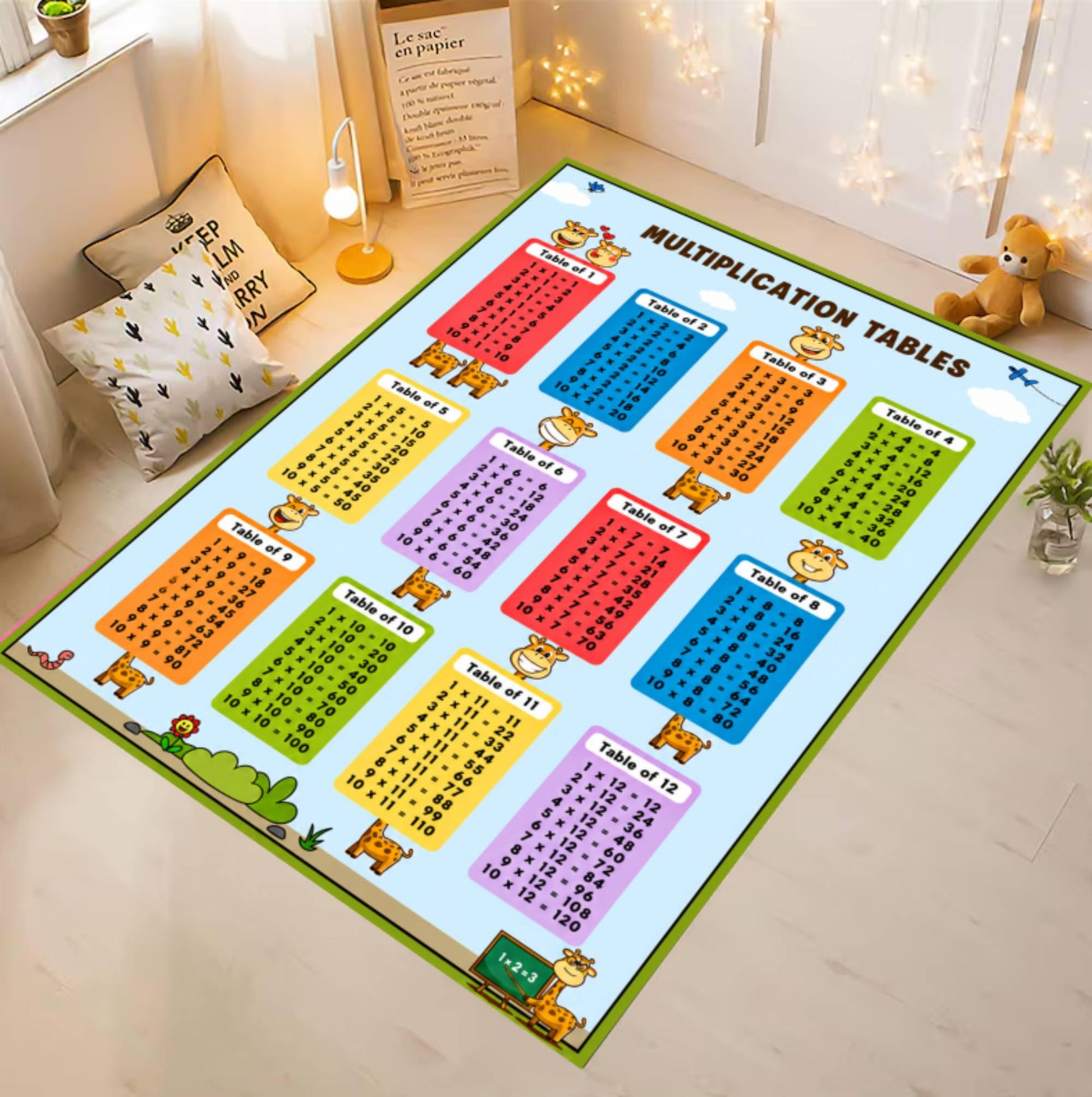 A large rectangular children's multiplication table mat laid out on a light wood floor in a styled room. The mat has a light blue background with multiple colorful tables (red, orange, yellow, green, blue, purple) and cartoon animal illustrations around the border.