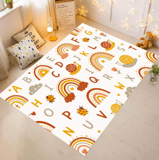 A rectangular children's play mat with a white background and colorful alphabet letters, rainbows, clouds, and sun motifs, shown laid out on a light wood floor in a styled room with pillows, a lamp, and string lights.