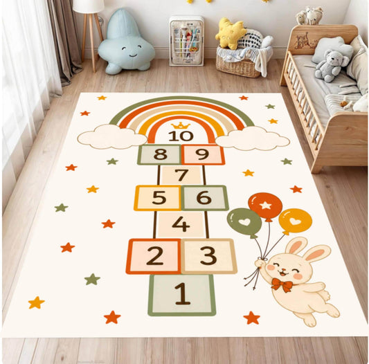 A rectangular children's play rug featuring a hopscotch grid with numbers 1 through 10, a rainbow, clouds, stars, and a cartoon bunny holding balloons, shown on a wooden floor in a styled nursery.